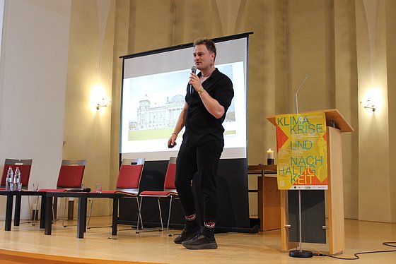 Joschi Wolf steht auf einer Bühne und redet in ein Mikrophon. Im Hintergrund ist eine Präsentation zu sehen, die das Reichstagsgebäude zeigt. 