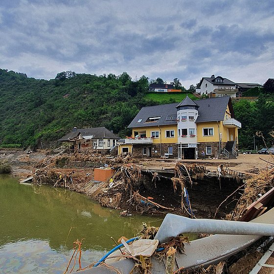 Im Vordergrund des Bildes ist ein trüber Fluss zu sehen, der hinten durch zerstörte Gebäudestrukturen und anderen Schutt begrenzt wird. Dahinter stehen zwei Gebäude, das eine ist stark zerstört. Im Hintergrund sind Himmel und Bäume zu sehen.