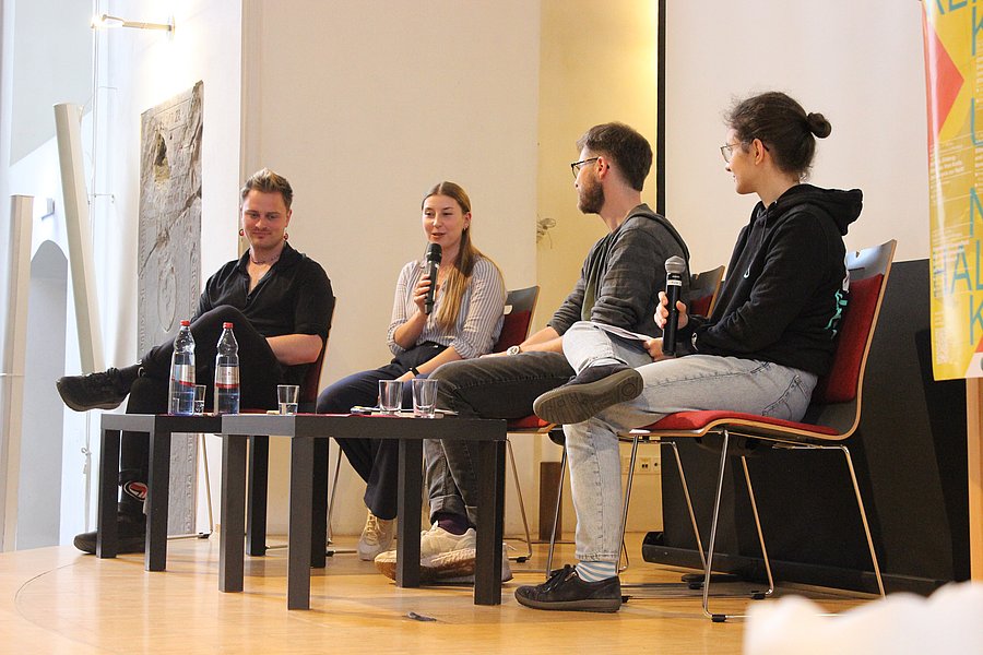 Vier Personen sitzen auf Stühlen auf einer Bühne. Von Links nach rechts: Joschi Wolf, Emilia Blank, Julius Merkens, Lea Hochkirchen. Emilia Blank spricht in ein Mikrophon.