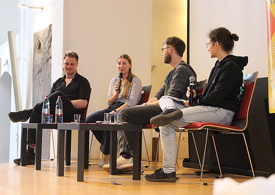 Vier Personen sitzen auf Stühlen auf einer Bühne. Von Links nach rechts: Joschi Wolf, Emilia Blank, Julius Merkens, Lea Hochkirchen. Emilia Blank spricht in ein Mikrophon.